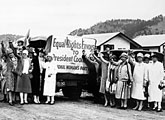 A group of women from the National Woman's Party stand beside a car with a sign reading, "Equal Rights Envoys to President Coolidge." They are outdoors, raising their right hands. Several houses and trees are visible in the background.