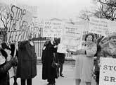 A black-and-white photo of a group of people protesting. They hold signs with various messages about opposition to the ERA (Equal Rights Amendment) and concerns about gender roles and the draft. The group is outside, and the atmosphere appears serious.