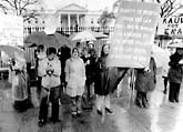A group of people stand in front of the White House holding umbrellas and signs advocating for the Equal Rights Amendment. One prominent sign displays the amendment's text. It is a rainy day, and the demonstrators wear coats. A group of people stand in front of the White House holding umbrellas and signs advocating for the Equal Rights Amendment. One prominent sign displays the amendment's text. It is a rainy day, and the demonstrators wear coats.